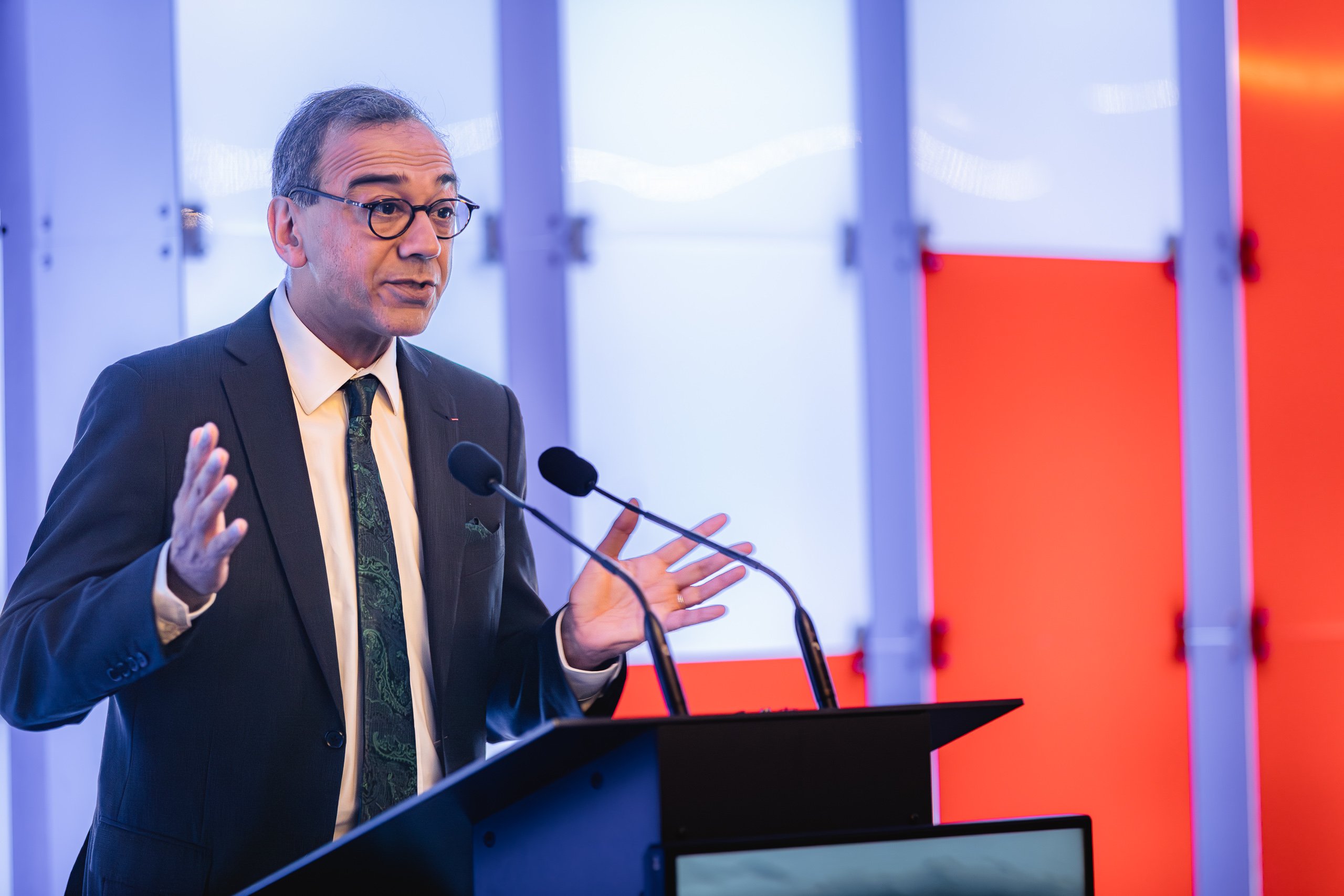 A man in a suit and glasses speaks at a lectern with two microphones. He gestures with his hands, appearing to address an audience. The background features panels lit in blue, white, and red tones, creating a modern, vibrant atmosphere.