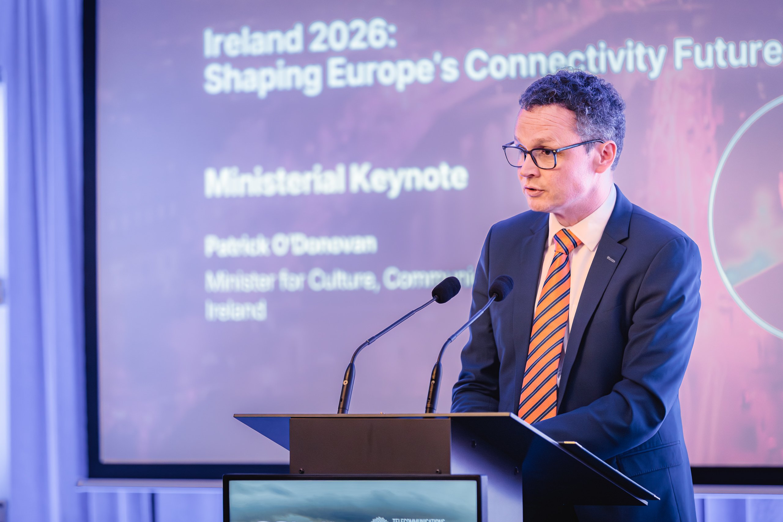 A man in a suit and orange-striped tie speaks at a lectern with two microphones. Behind him, a large screen displays the event title: "Ireland 2026: Shaping Europe’s Connectivity Future" and his role as Minister for Culture, Communications, Ireland.