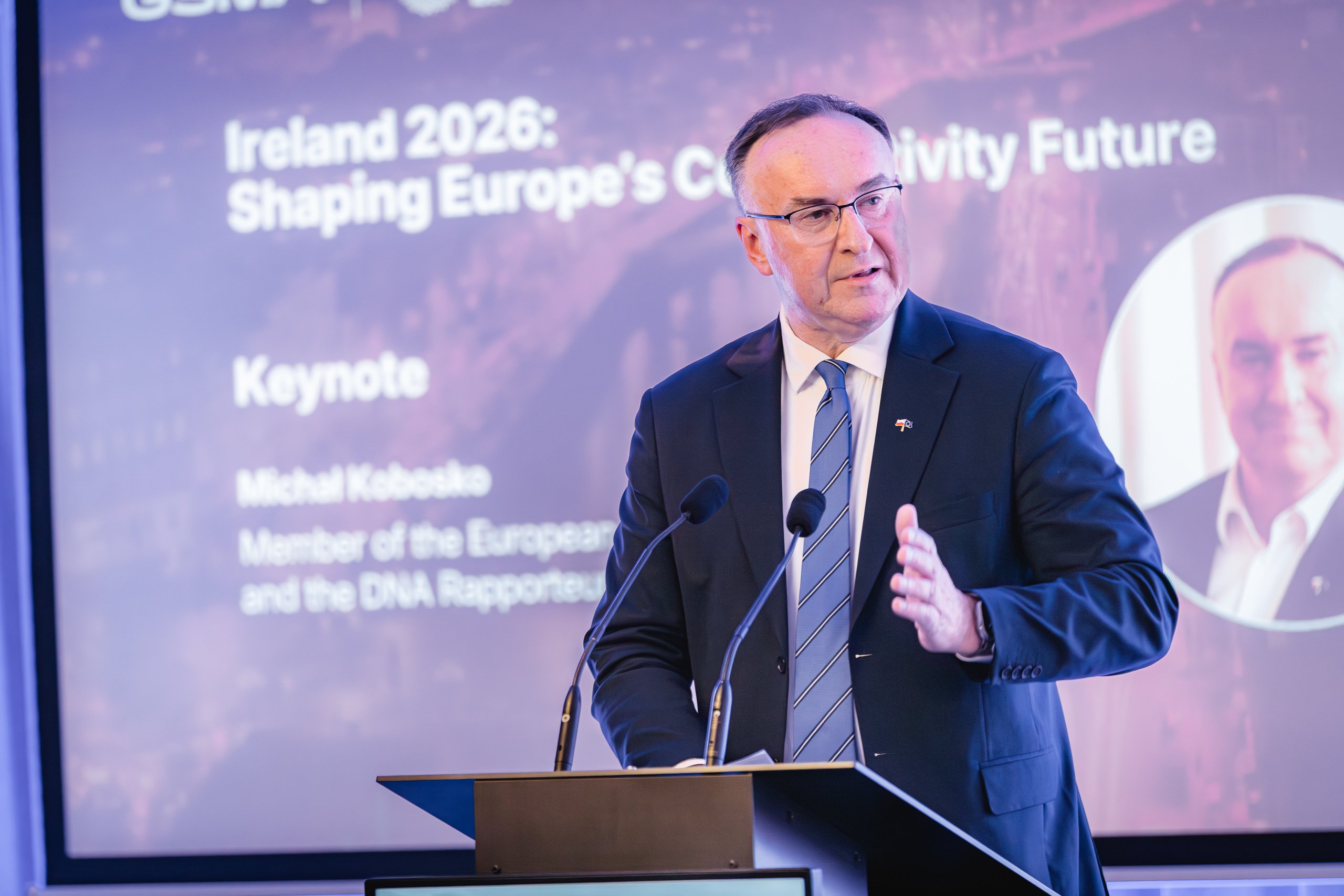 A man in a dark suit, tie, and glasses speaks at a lectern. Behind him is a large screen displaying text for a keynote: “Ireland 2026: Shaping Europe’s Connectivity Future” and the speaker’s name, Michał Kobosko, with his photo inset on the right.