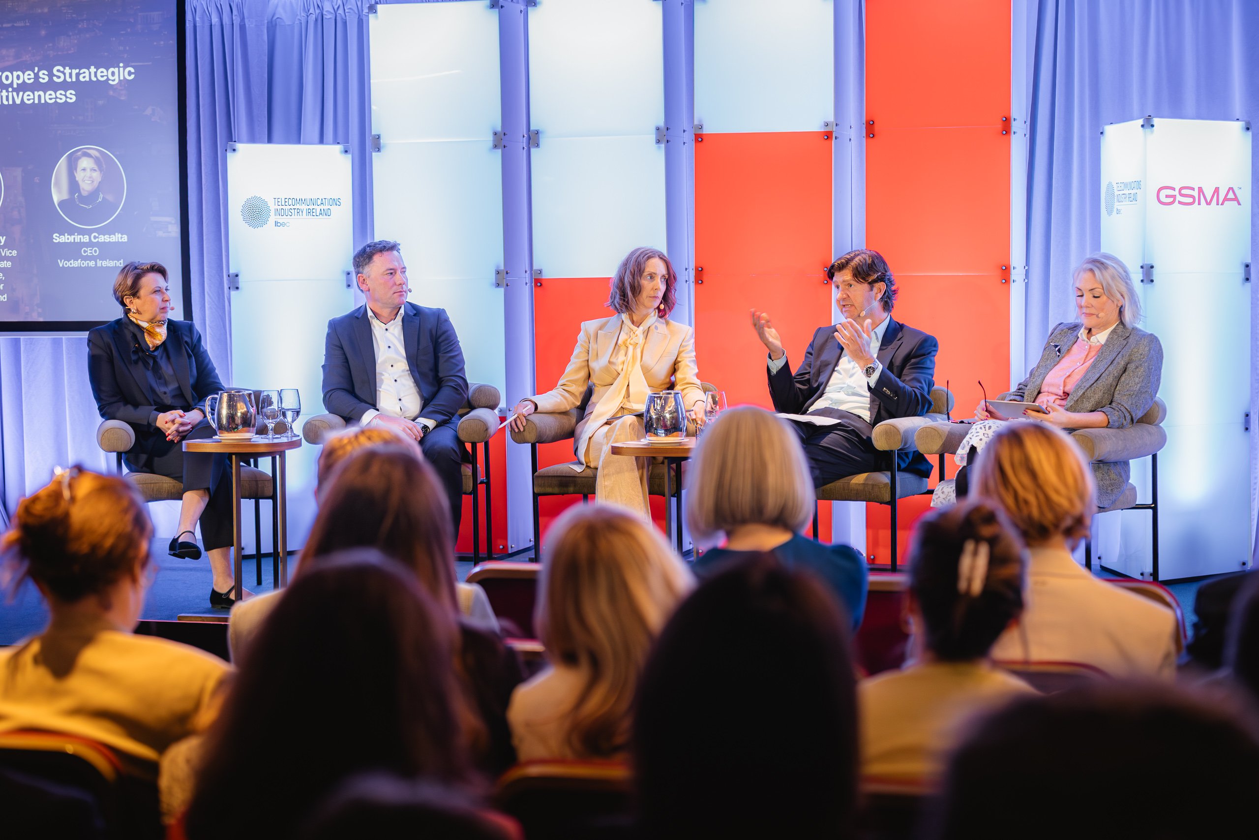 Five panellists sit on stage, engaged in discussion at a conference. Three women and two men face an audience. Bright lights, a backdrop with coloured panels, and event logos are visible. One man gestures while speaking; others listen attentively.