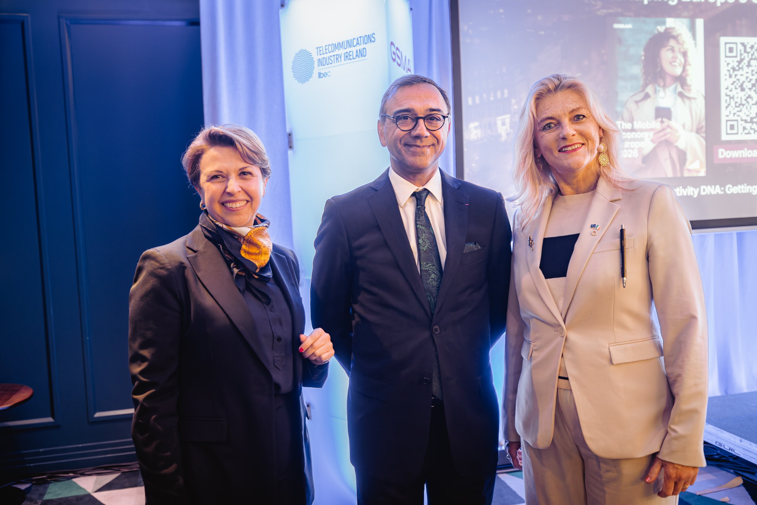 Three people stand together indoors, smiling at the camera. Two women wear suits—one in black with a scarf, the other in white—whilst the man in the centre wears a dark suit and glasses. Behind them is event signage with telecommunications branding.