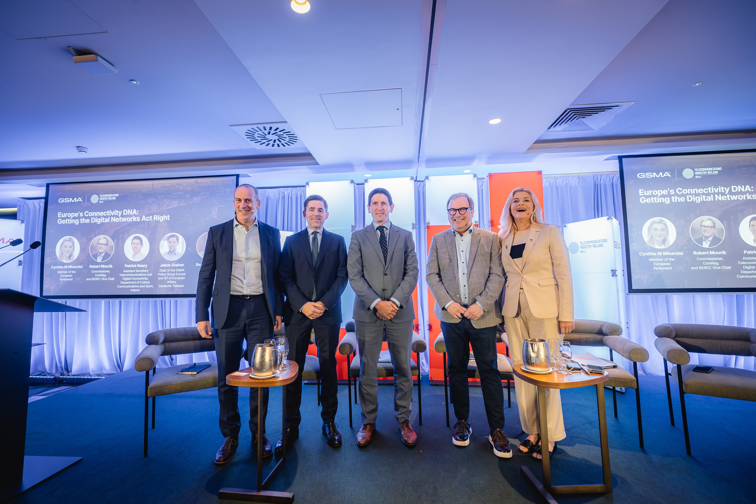 Five people in business attire stand smiling on a stage with blue lighting. Behind them are chairs, tables with jugs, and two screens displaying event info titled “Europe’s Connectivity DNA: Getting the Digital Networks Act Right” with panellist names and photos.