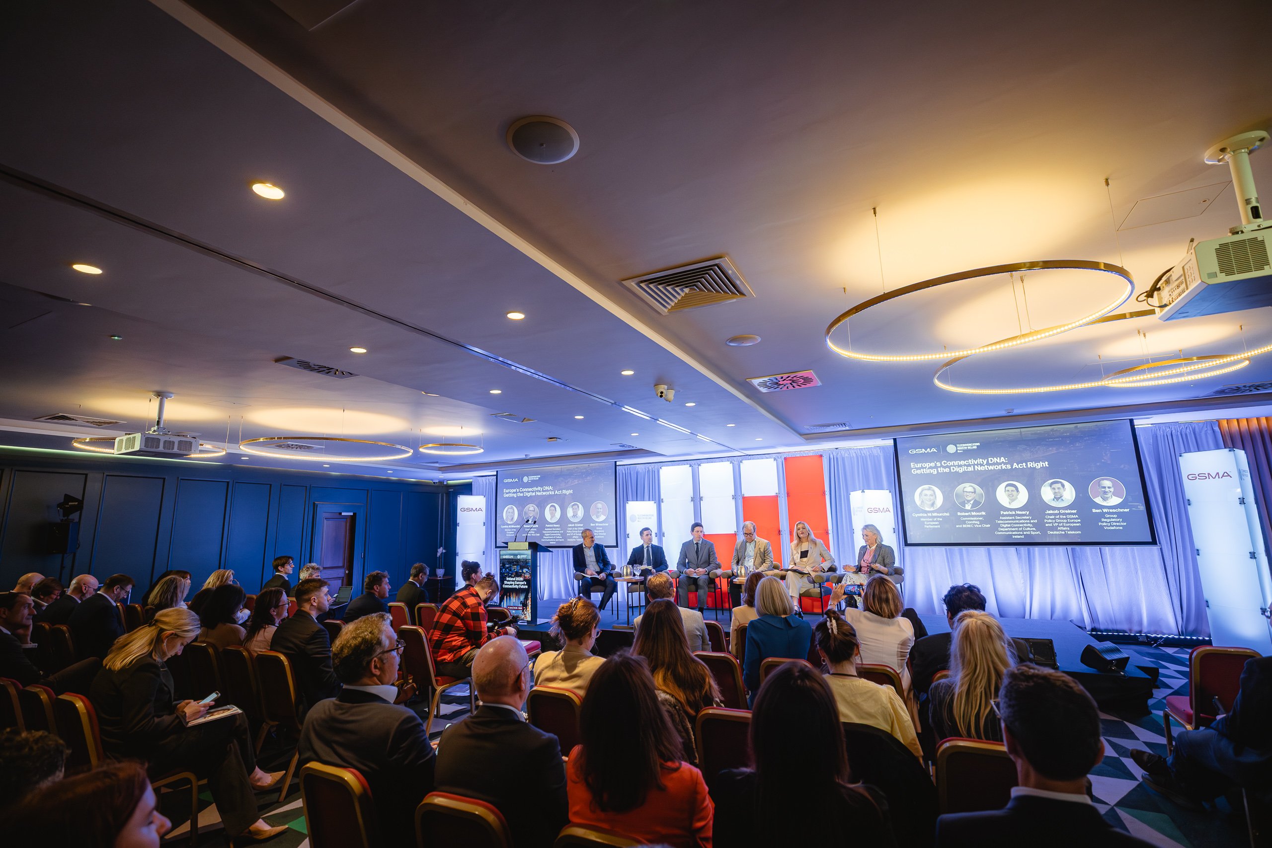 A conference room with an audience seated facing a panel of five speakers on a stage. Large screens behind the panel display speaker names and topics. The room is brightly lit with circular ceiling lights and has a formal, professional atmosphere.