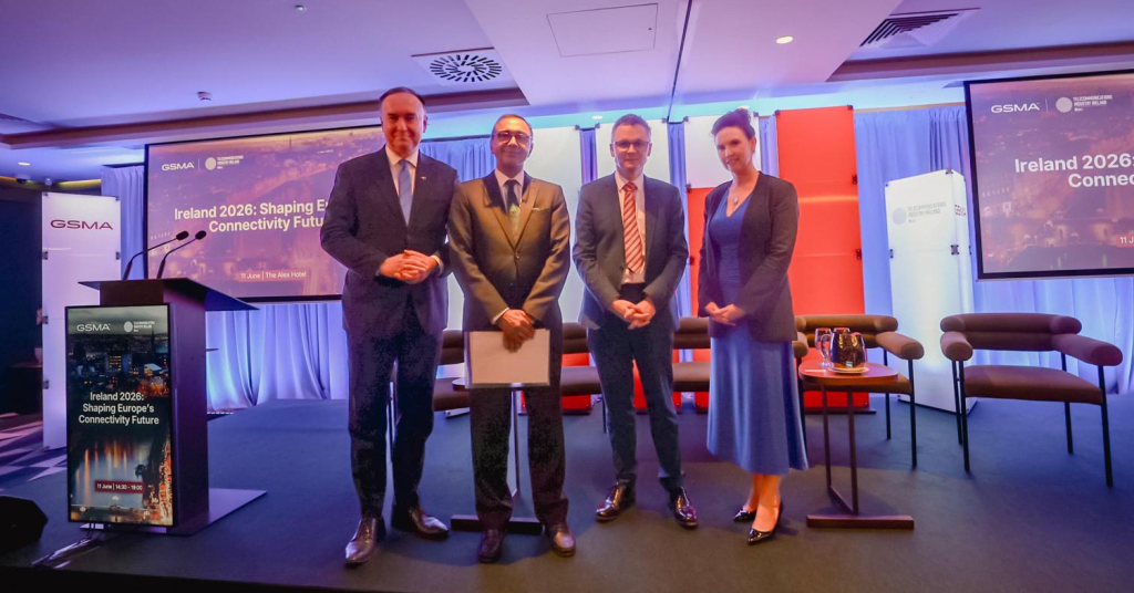 Four people in business attire stand on a stage, posing and smiling for a photo. Behind them is a screen displaying “Ireland 2026: Shaping Europe’s Connectivity Future” with logos and colourful lighting. Chairs and a small table are arranged nearby.