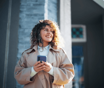 A woman with curly hair, wearing a beige trench coat and white polo neck, stands outside holding a mobile phone in both hands. She smiles and looks off to the side. The background features a grey stone wall and a blurred window.