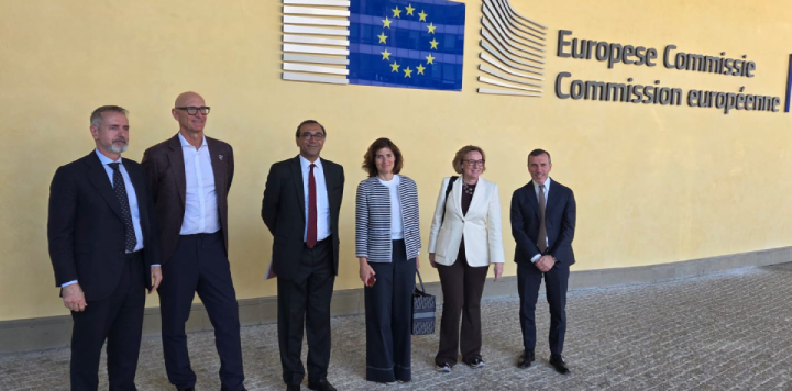 Six people in business attire stand in a row outside a building with a large European Union flag and “European Commission” written in English and French on the wall behind them. They are posing for a group photo on a paved surface.