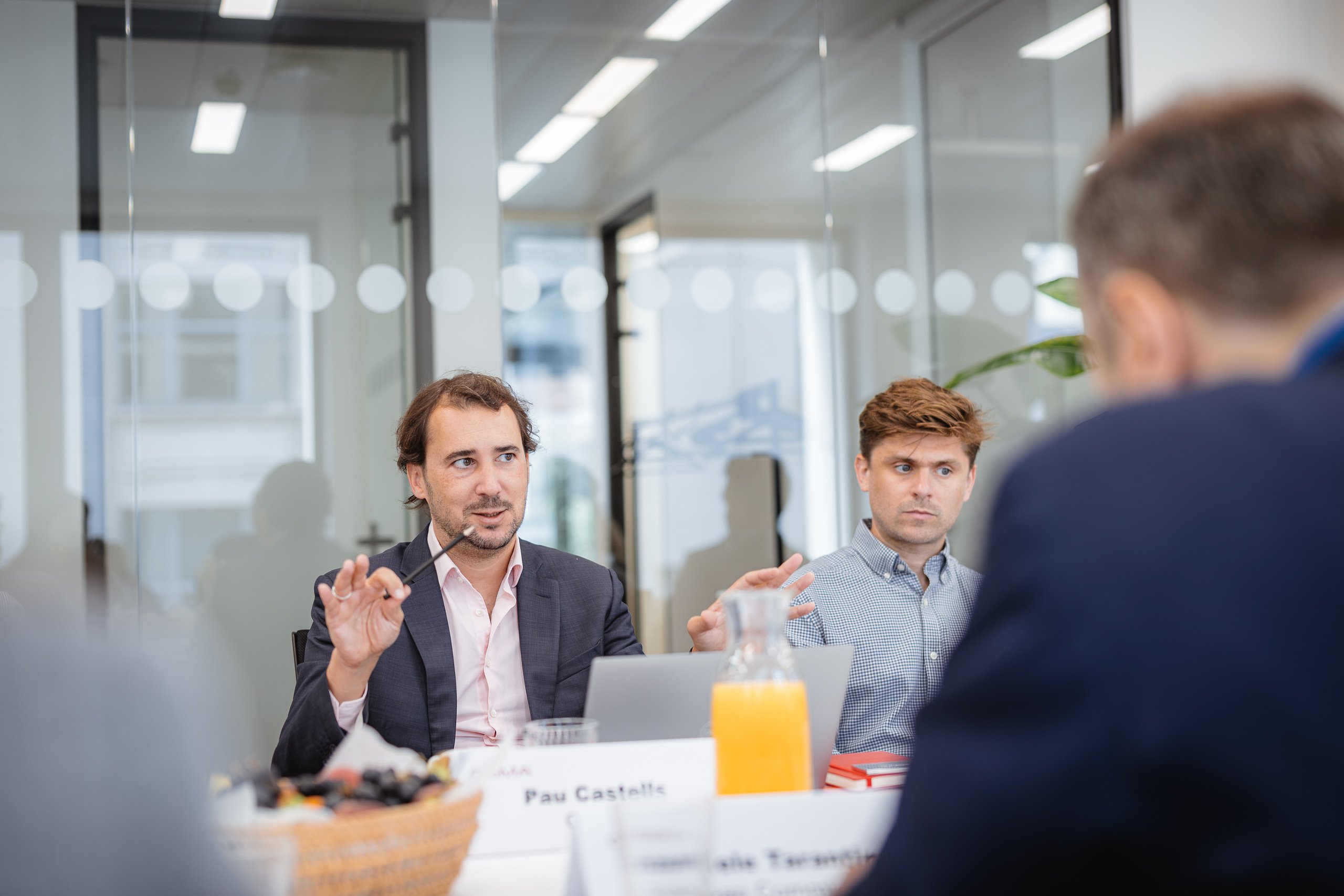 A man in a suit gestures while speaking at a meeting table with another man. Nameplates, juice, and fruit are on the table. Glass walls reflect office lights, and two blurred figures are in the foreground. The scene appears professional and collaborative.