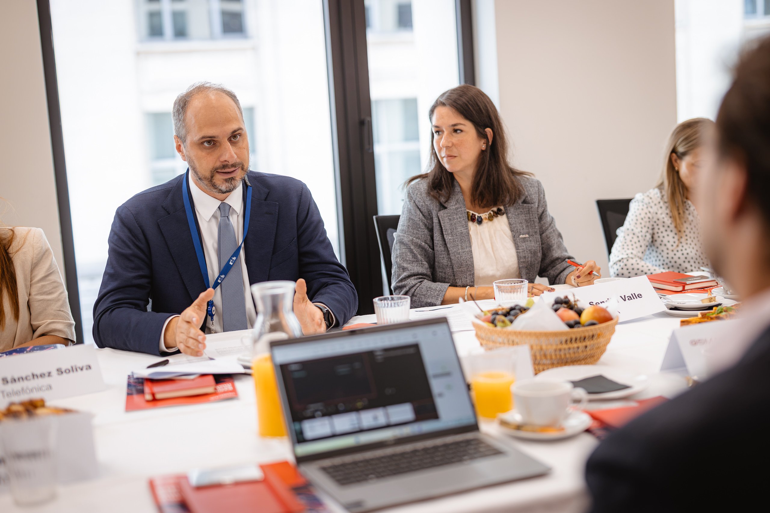 Four people sit around a conference table with documents, fruit, and drinks. A man in a suit gestures while speaking; a woman beside him listens. A laptop, paperwork, and juice are visible. Large windows show a city view in the background.