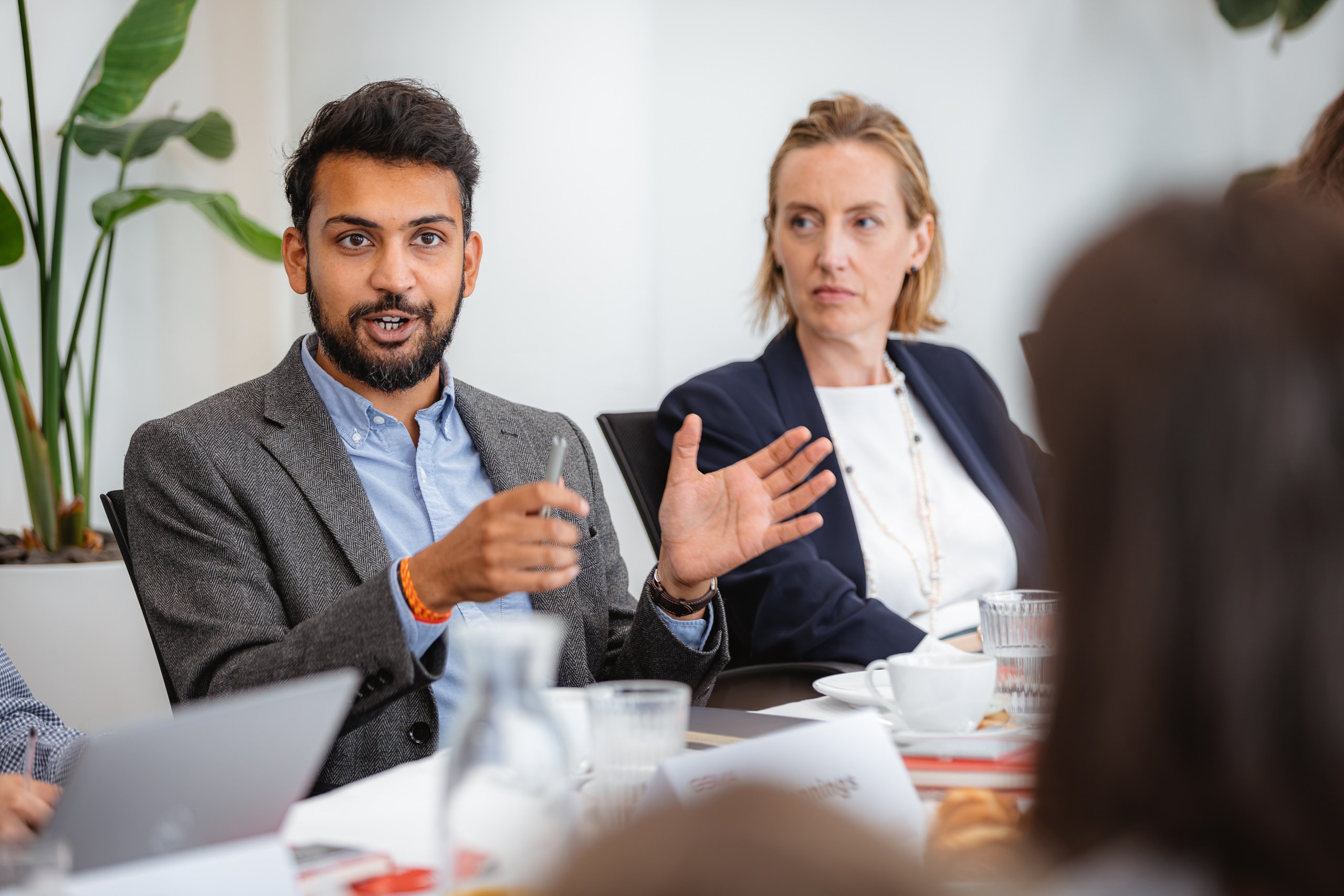 A man with a beard in a grey blazer gestures while speaking at a meeting table, with a woman in a navy jacket listening beside him. Papers, glasses of water, and a laptop are visible; plants and white walls are in the background.