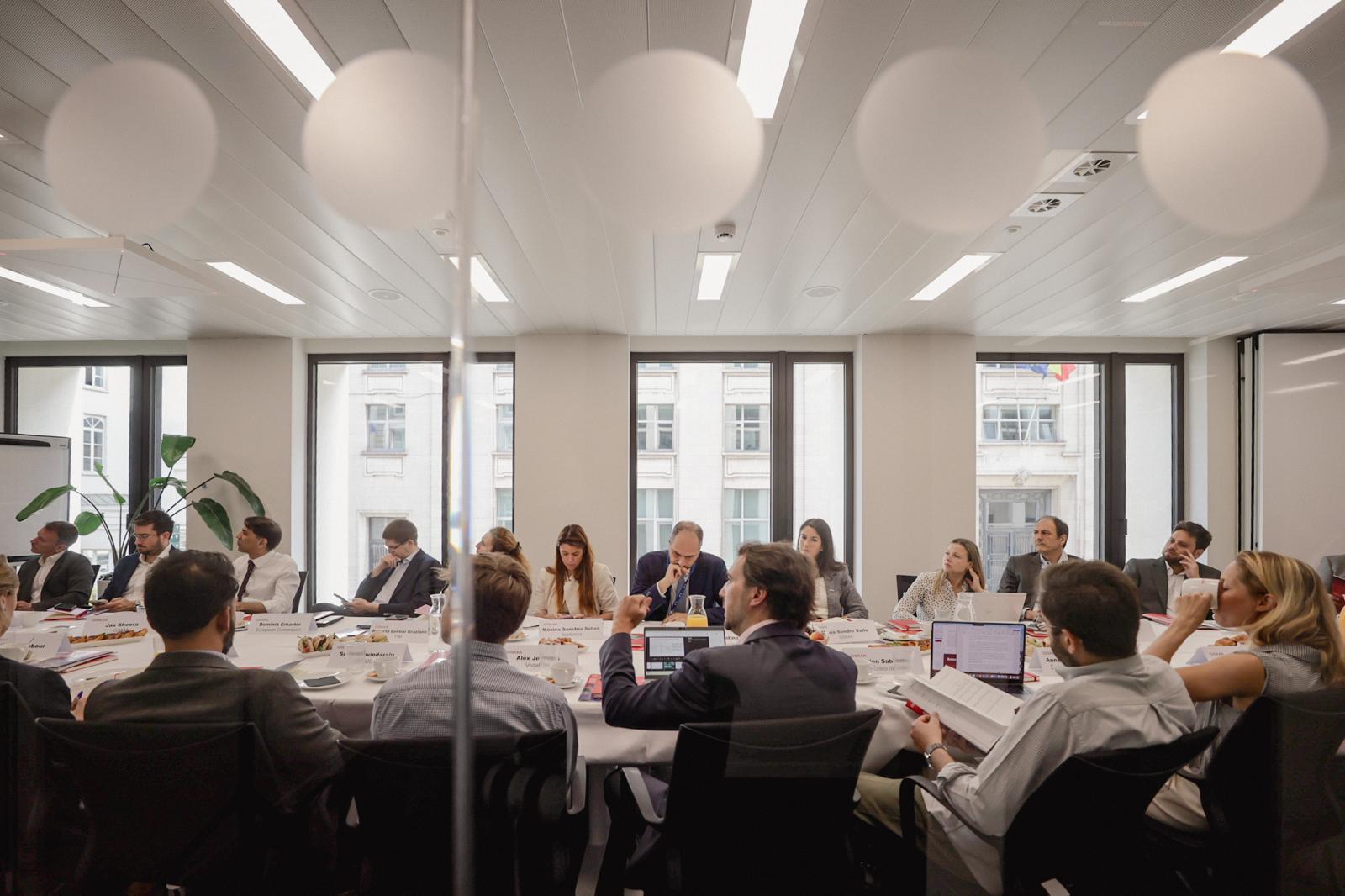 A group of people in business attire are seated around a large oval table in a modern office meeting room, engaged in discussion. Laptops, papers, and food are on the table. Large windows line the back wall, letting in natural light.