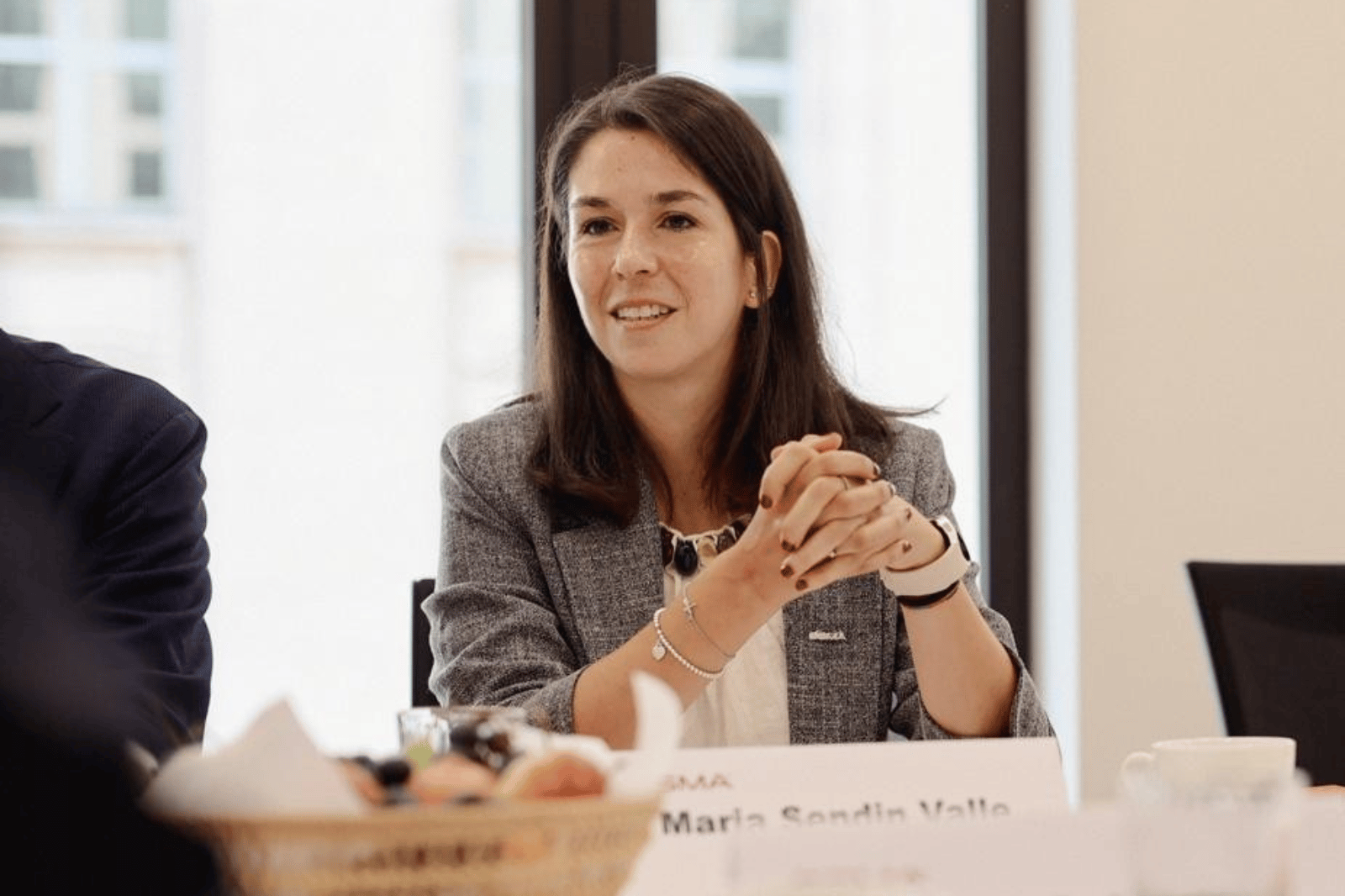 A woman with long brown hair, wearing a grey blazer and white top, sits at a table with her hands clasped. There is a blurred fruit bowl and a nameplate partially reading "Maria Sandin Valle" in front of her, and a window in the background.