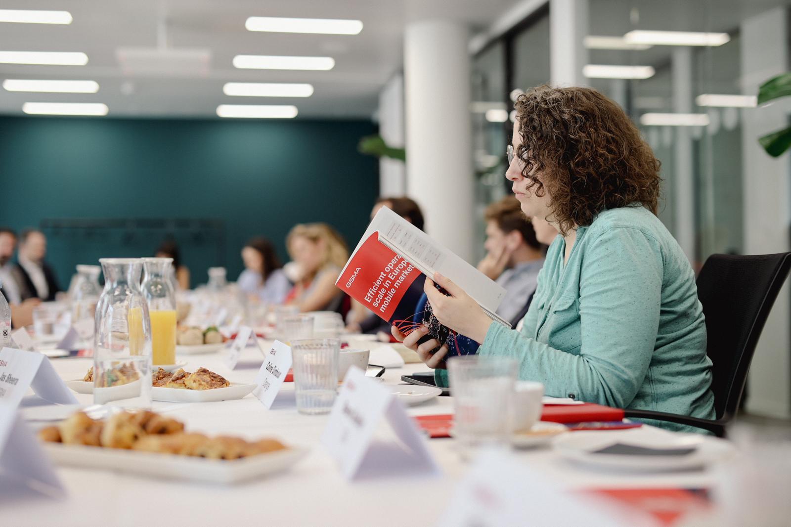 A woman with curly hair and glasses sits at a conference table reading a red-covered book. The table is set with pastries, juice, glassware, and name badges. Other attendees are present in a modern office setting with large windows and teal walls.