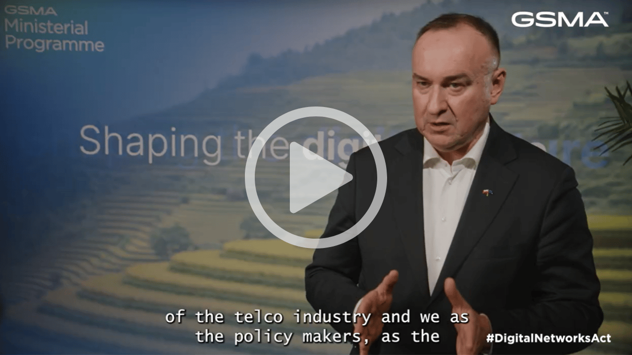 A man in a dark suit gestures whilst speaking in front of a scenic backdrop with the text “Shaping the digi” partially visible. GSMA and “Ministerial Programme” appear at the top. Subtitles and hashtag #DigitalNetworks are displayed at the bottom. A video play button overlays the image.