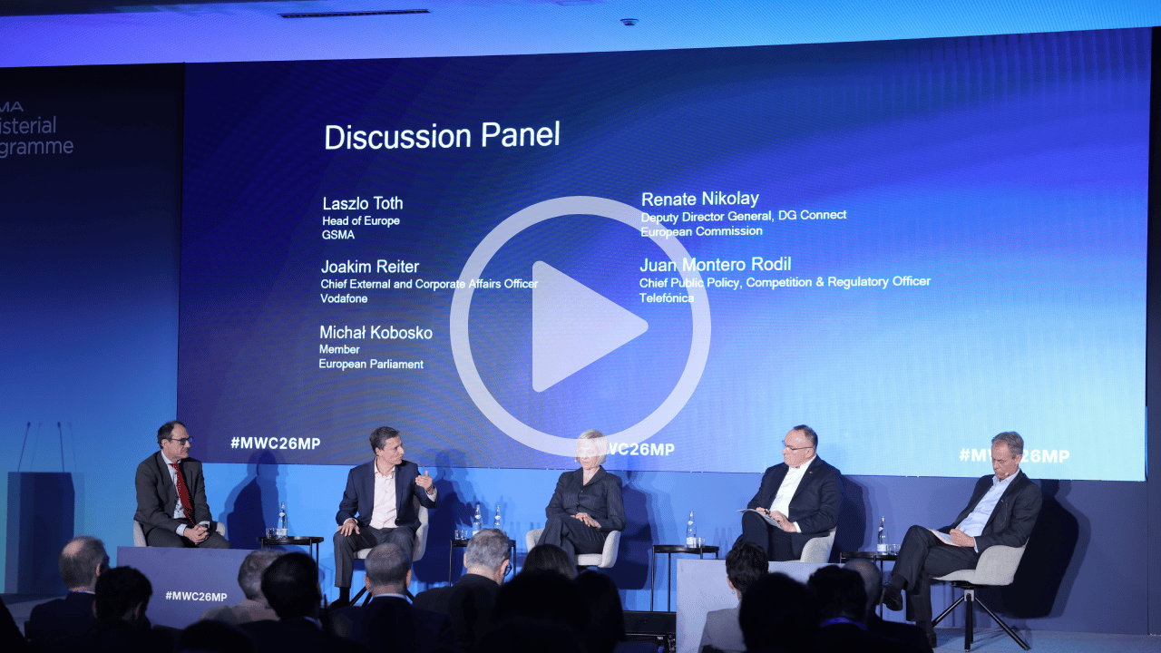 Five people sit on stage during a panel discussion. Behind them, a blue screen displays their names and titles. There are chairs, small tables with water bottles, and microphones. The audience is not visible. A large play button is centred on the image.
