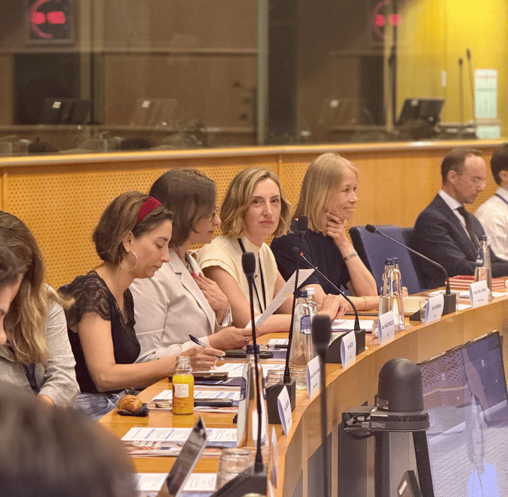 A group of professionals, mostly women, sit at a conference table with microphones, documents, bottled water, and nameplates. They are engaged in discussion in a well-lit meeting room with wood panelling and glass dividers.