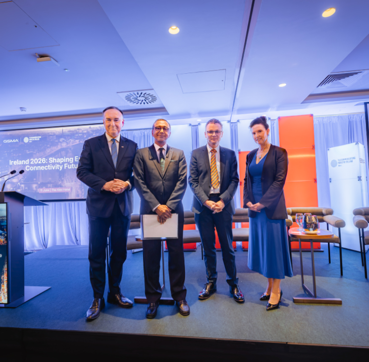 Four formally dressed people stand on a stage in front of a screen and chairs at a conference. There are two men in suits on the left, one man in a suit and one woman in a blue dress on the right. The background features banners and curtains.