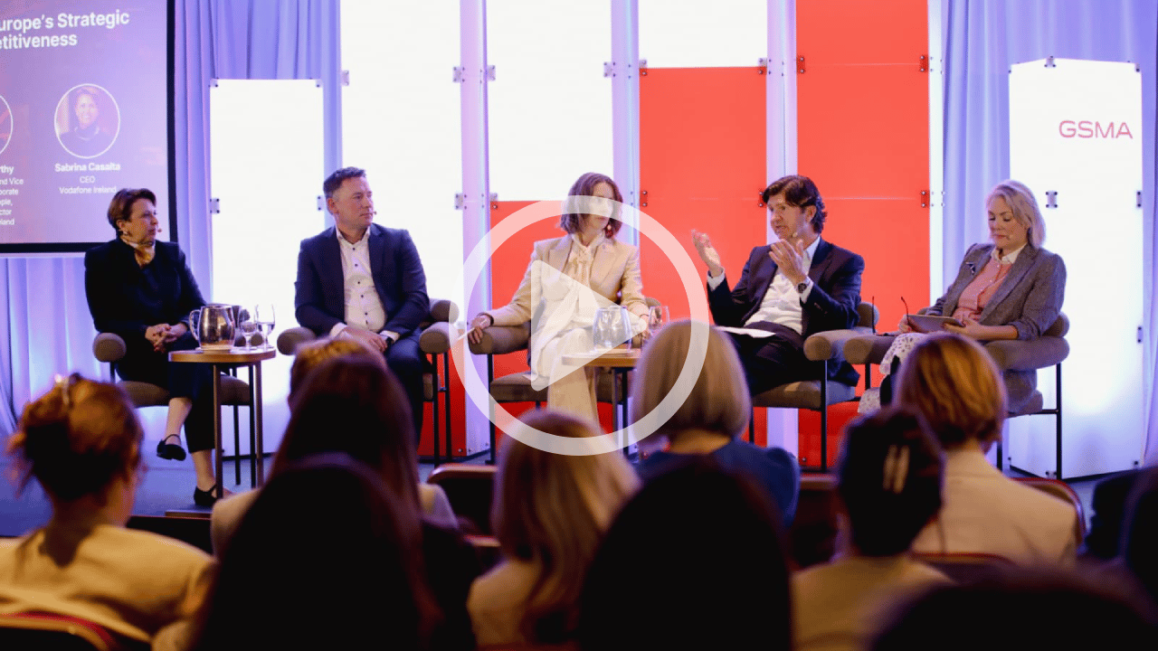 Five people sit onstage in chairs, speaking at a panel discussion. An audience watches them. Behind the panellists are illuminated screens, one with the text “GSMA.” Drinks and notepads are on a table. A translucent play button icon overlays the image.