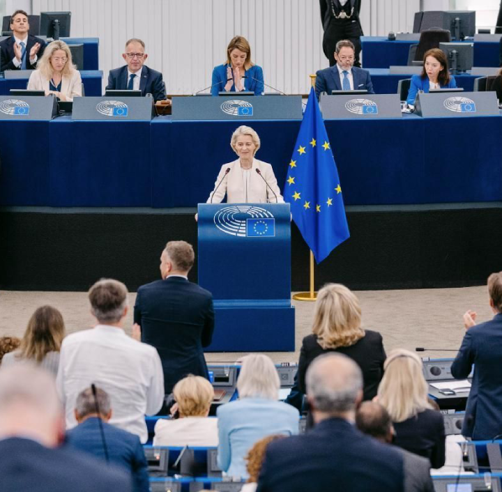 Ursula von der Leyen stands at a lectern in the European Parliament, addressing a chamber of seated members. She wears a cream-coloured blazer, with a large European Union flag positioned to her right. Behind her, other officials are seated at a long desk in front of blue panels.