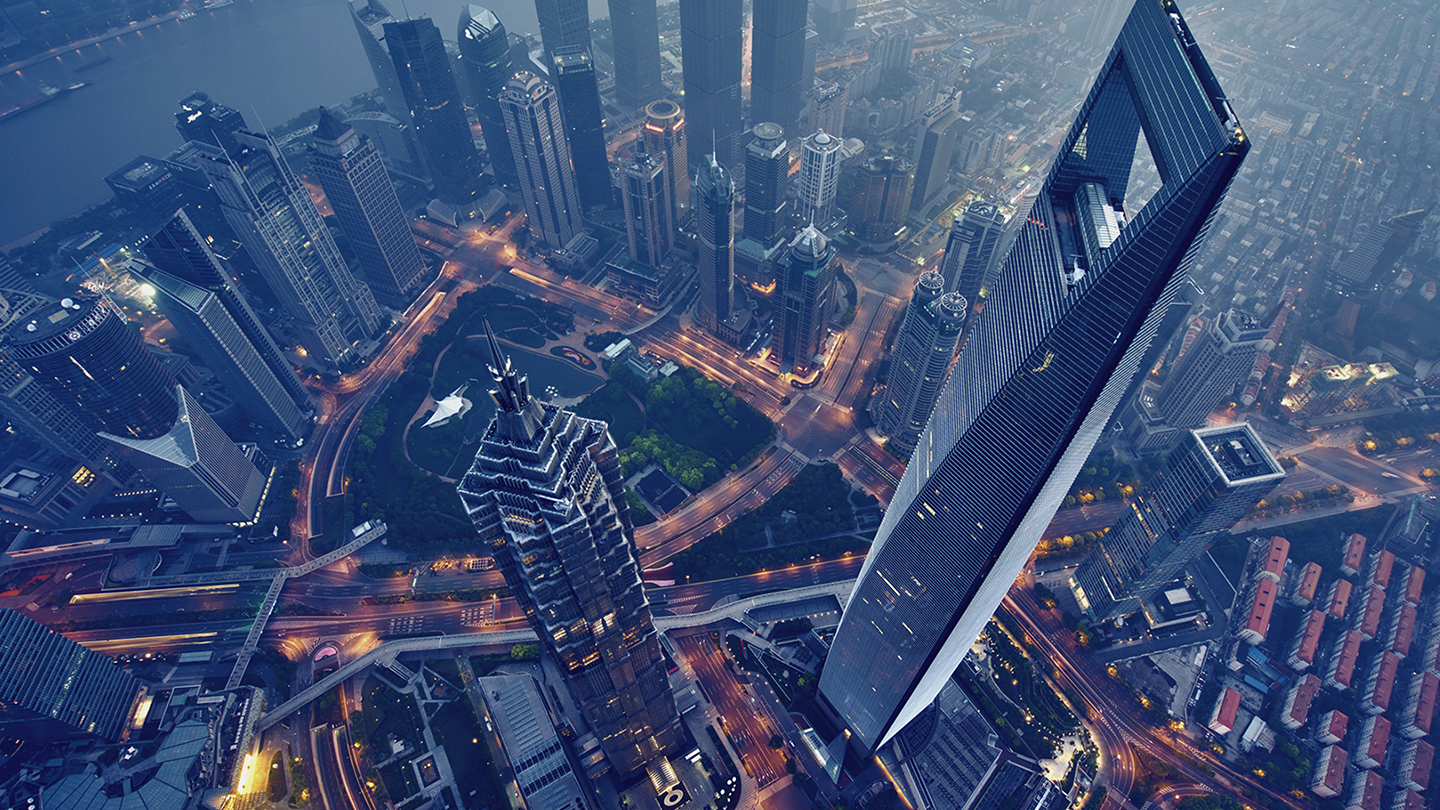 Aerial view of a densely built-up cityscape featuring numerous high-rise buildings, including the distinctively shaped Shanghai World Financial Center. Streets with vehicular traffic and illuminated intersections are visible amidst the urban setting.