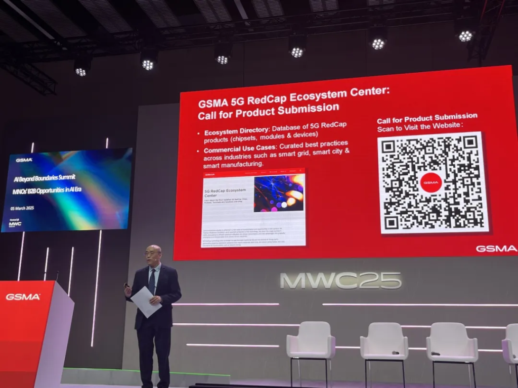 A man in a suit stands on stage holding papers. Behind him is a large screen displaying a red slide titled "GSMA 5G RedCap Ecosystem Center." It features a QR code and text about product submissions. Three white chairs are lined up on the stage, and a colorful side screen reads "MWC23.