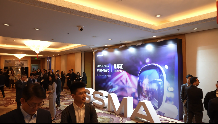 A group of people in business attire mingle in a hotel lobby at an event. A large illuminated display reads “2023 GSMA Post-MWC” in English and Chinese, alongside a graphic of a smartphone and a woman’s face. Large GSMA letters are positioned on the floor.