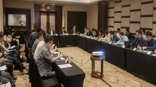 A large group of people in business attire are seated around three sides of a rectangular arrangement of tables in a conference room, engaged in a meeting. Papers, nameplates, and a projector are visible on the tables. The atmosphere appears formal and focused.