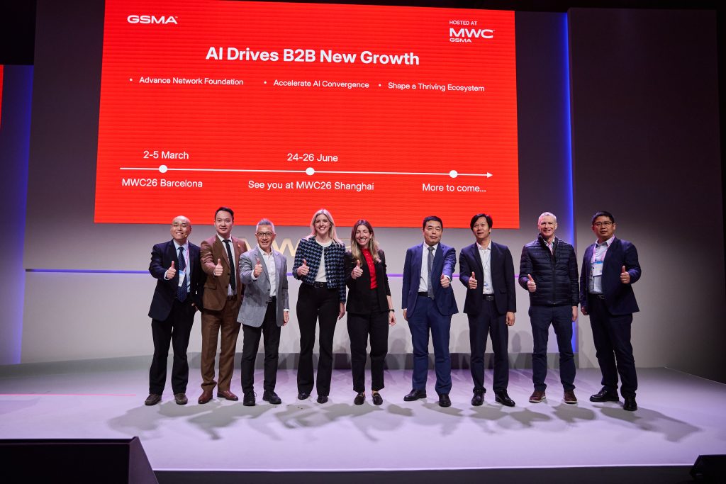 Nine people stand on stage giving thumbs-up in front of a red screen at an AI business event.