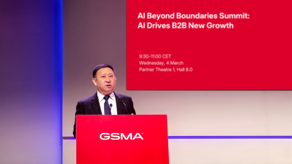 A man speaks at a podium during the "AI Beyond Boundaries Summit" with a red event screen behind him.