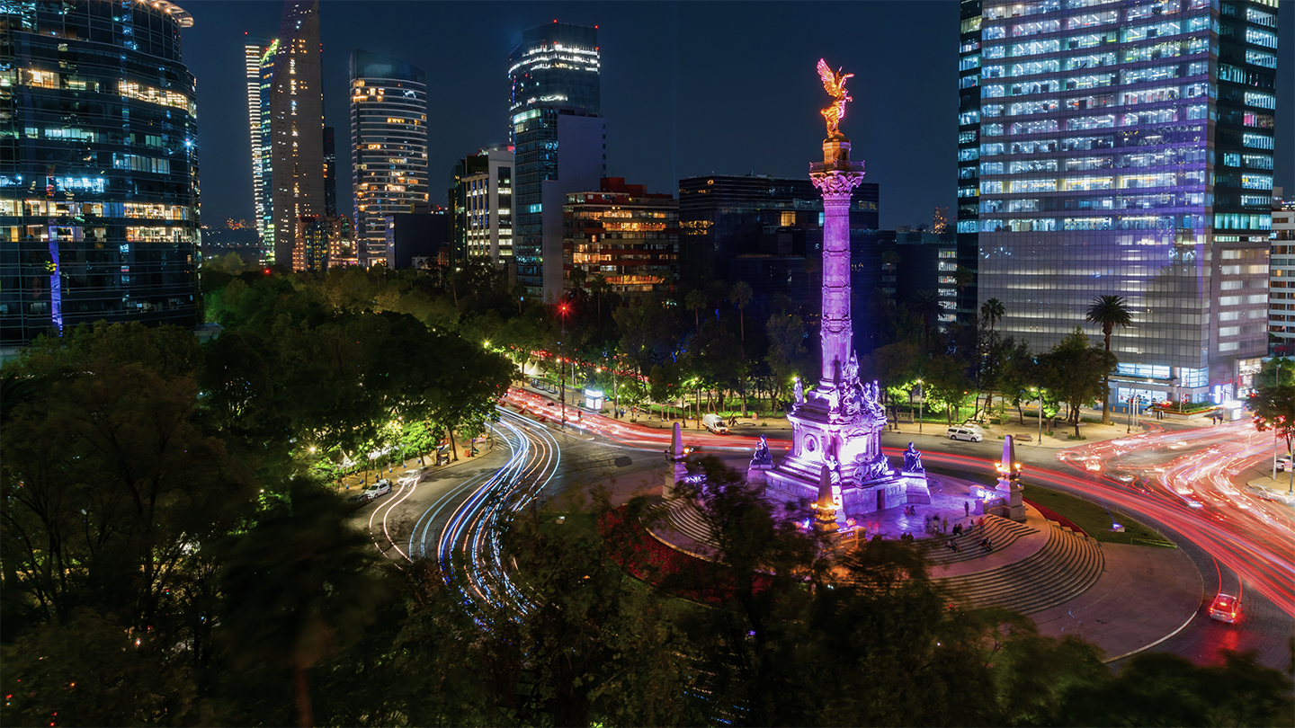 A brightly lit roundabout at night, featuring a large monument with a golden angel on top, surrounded by tall, illuminated modern buildings. Light trails from cars indicate traffic movement around the roundabout. Green foliage fills the lower part of the scene.