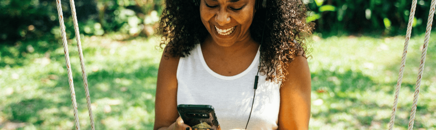 Una mujer de cabello rizado está sentada en un columpio al aire libre, con una camiseta blanca sin mangas y auriculares. Sonríe y mira su teléfono. El fondo muestra vegetación con árboles y plantas difuminados.