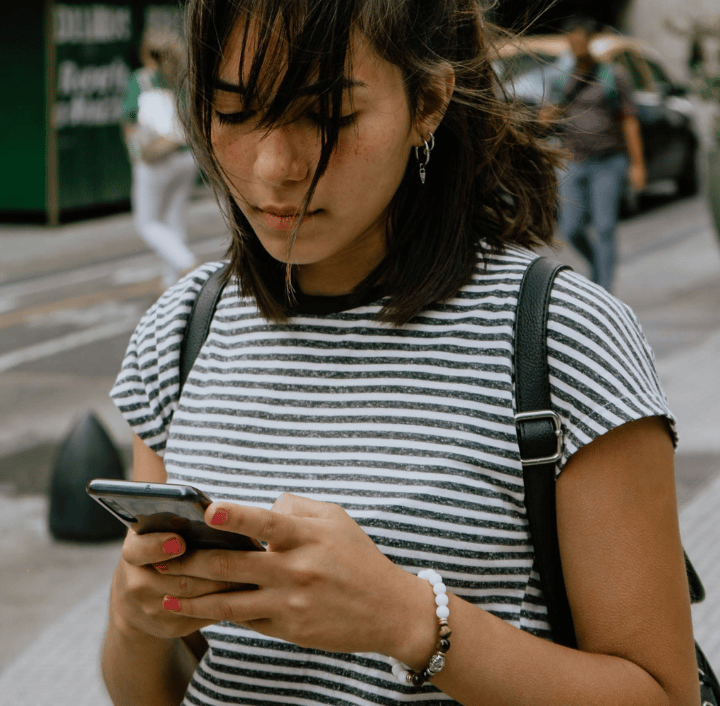 Una mujer joven con el pelo hasta los hombros mira su teléfono mientras está parada en una calle de la ciudad. Lleva una camiseta de rayas blancas y negras y una pulsera de cuentas, y lleva una mochila negra sobre los hombros. Hay personas y vehículos en el fondo.