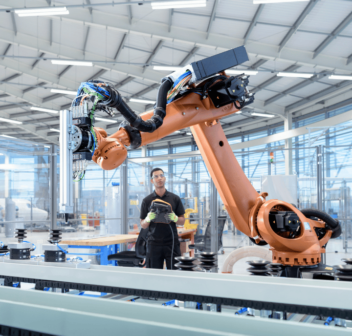 Una persona se encuentra en una fábrica moderna, sosteniendo una tableta digital y mirando un gran brazo robótico industrial de color naranja. El brazo robótico está operando cerca de una línea de producción bajo un techo alto con tragaluces, rodeado de varias máquinas y equipos.