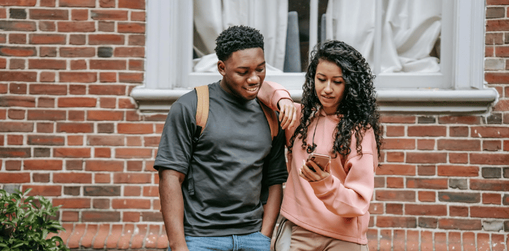 Two people stand in front of a brick wall. The person on the left is smiling and has a backpack. The person on the right is wearing a pink sweatshirt and looking at a smartphone. A window with white curtains is in the background.