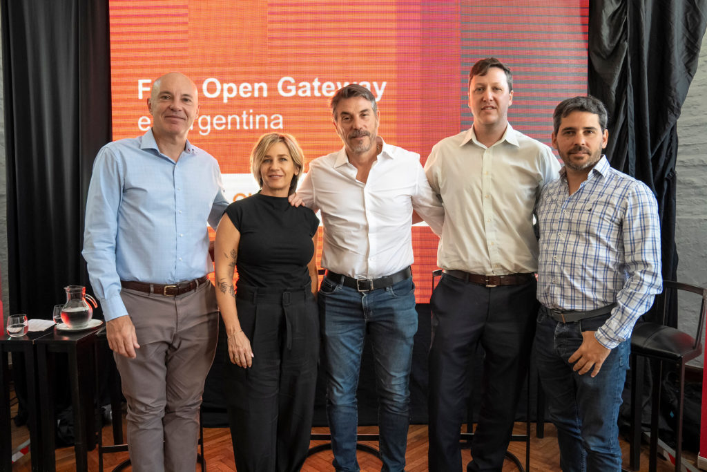 Five people pose together in front of a red and orange backdrop with the text "Open Gateway Argentina." Standing indoors on a wooden floor, dressed in business casual attire, the group includes four men and one woman, all smiling warmly at the camera.