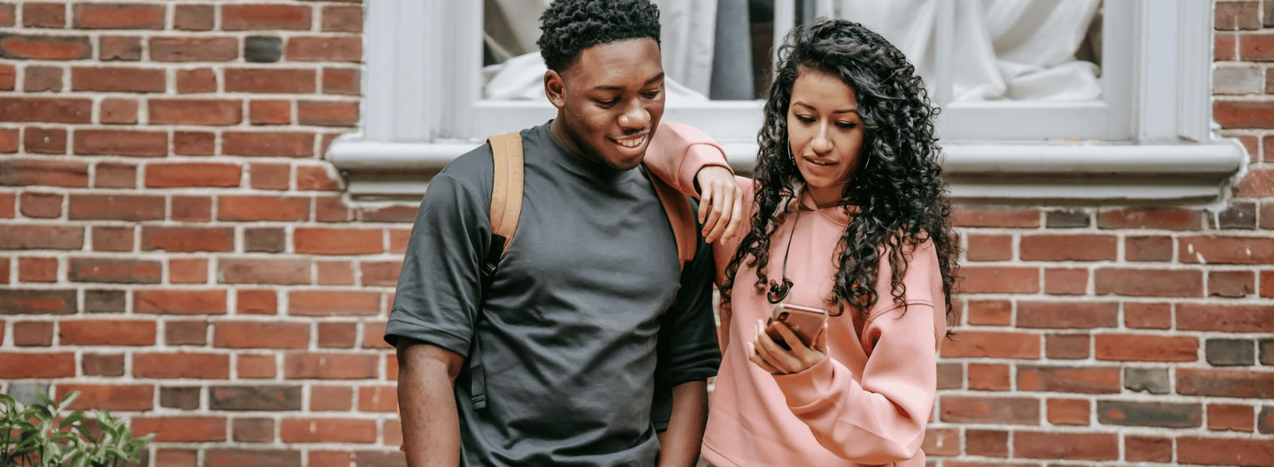 A young man and woman stand in front of a brick wall. The man, wearing a black shirt and backpack, looks at the phone the woman is holding. The woman is wearing a pink hoodie. They are outside, near a window with a white curtain.