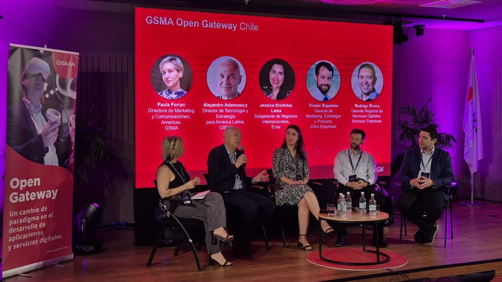 At the "GSMA Open Gateway Chile" event, a panel of five people sits on stage in front of a large red screen displaying their photos and names. Nearby, a banner reads "Open Gateway." The dimly lit room is awash with purple lights, while a Japanese flag subtly adorns the background.