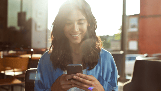 A woman with long hair, wearing a blue shirt, smiles while looking at her smartphone. She is seated indoors, with sunlight streaming through large windows, creating a warm and bright atmosphere. The background shows blurred chairs and part of a building outside.