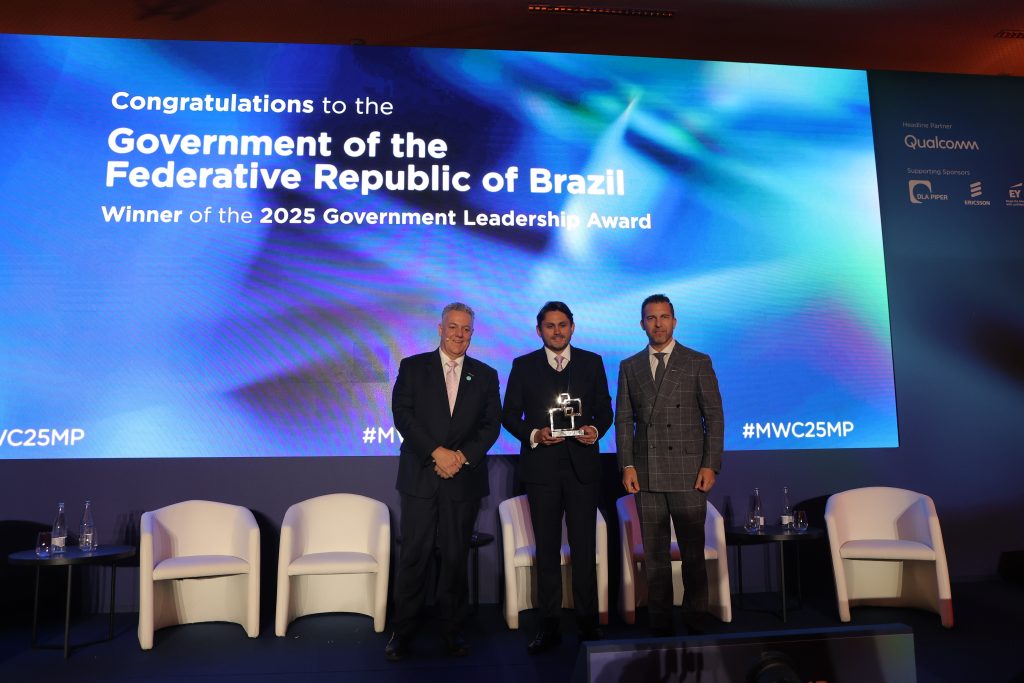 Three men in suits stand on stage at the Brasil Prêmio Government Leadership Award ceremony for the Federative Republic of Brazil 2025. One man holds a trophy, while a large screen behind them displays event details. Empty chairs are positioned alongside them.