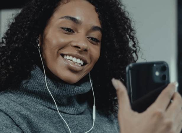 A smiling woman with curly hair, wearing a gray turtleneck sweater and wired earphones, holds a smartphone in her hand and looks at the screen, appearing to enjoy a video call or conversation.