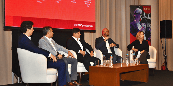 Cinco personas en traje de negocios se sientan en un panel del escenario, enfrascadas en un debate. Cuatro hombres y una mujer sostienen micrófonos, con una gran pantalla roja detrás de ellos que muestra el logotipo de la GSMA y Open Gateway Paraguay. A un lado hay un cartel con un casco de realidad virtual.