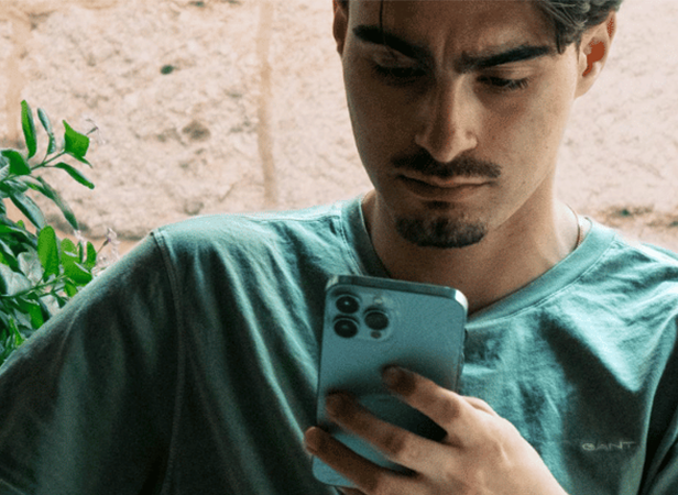 Un joven de pelo castaño corto y barba recortada, vestido con una camiseta verde, mira atentamente un smartphone que tiene en la mano. Al fondo, una frondosa planta verde y una pared beige con textura. Parece concentrado o ligeramente preocupado.