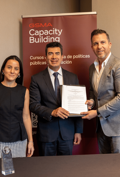 Tres personas sonríen junto a una mesa; dos hombres sostienen un documento delante de una pancarta de "GSMA Capacity Building".
