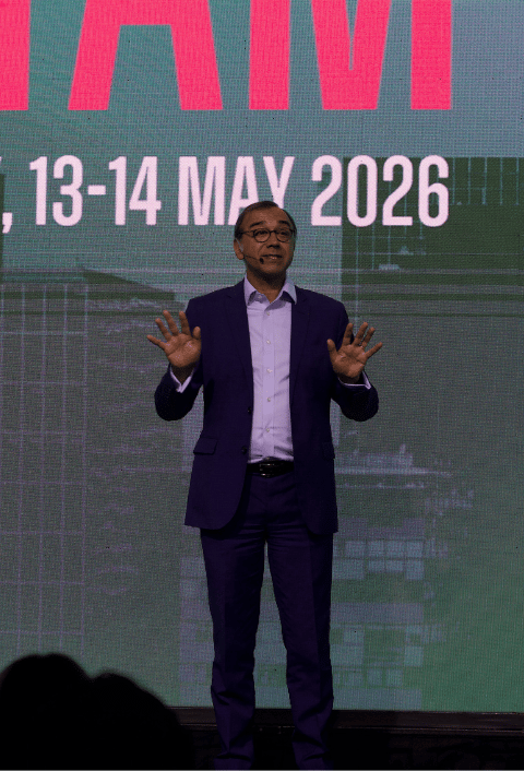 Un hombre trajeado habla en el escenario con el mensaje "13-14 de mayo de 2026" en la pantalla detrás de él.