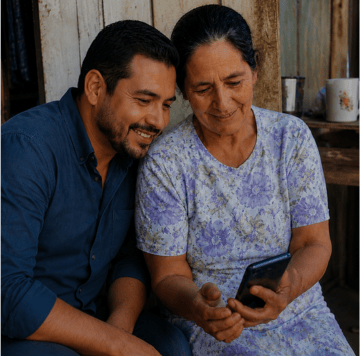 Hombre sonriente y mujer mayor sentados juntos, mirando un smartphone al aire libre cerca de una pared rústica de madera.