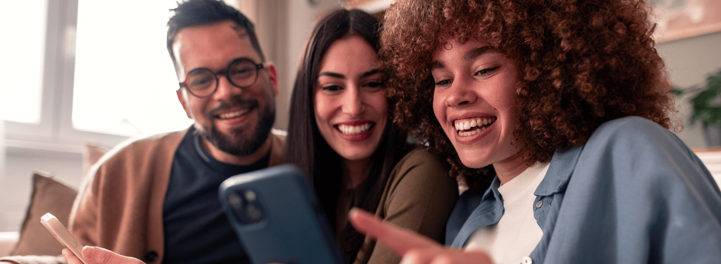 Tres amigos sentados juntos en un sofá, sonriendo y mirando un smartphone.