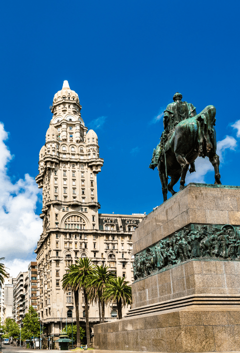 Un edificio alto y ornamentado, coronado por una cúpula, se alza bajo un cielo azul. En primer plano se ve una estatua ecuestre de bronce sobre un gran pedestal de piedra, rodeada de palmeras y edificios de la ciudad. La escena es animada y está bañada por la luz del sol.