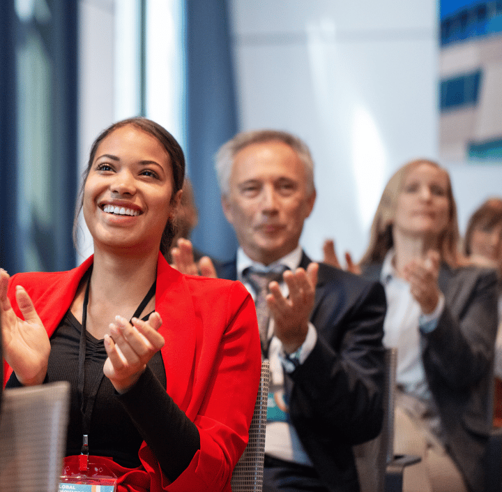Una mujer con una chaqueta roja sonríe y aplaude en un acto. Detrás de ella, varios profesionales del mundo empresarial también aplauden. El escenario es una moderna sala de conferencias con abundante luz natural y detalles difuminados en el fondo.