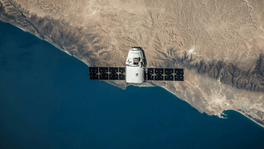 Un satélite blanco con paneles solares negros orbita sobre una costa escarpada, mostrando claramente la tierra y el mar de un azul intenso que se extiende a sus pies, con montañas y terrenos desérticos visibles a lo largo de la costa.