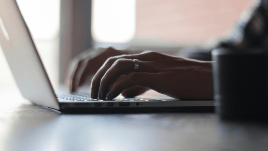 Primer plano de una persona escribiendo en el teclado de un ordenador portátil. La persona lleva un anillo en la mano izquierda. El fondo está ligeramente desenfocado, con la luz del día entrando por una ventana. En primer plano se ve parcialmente un objeto cilíndrico oscuro y borroso.