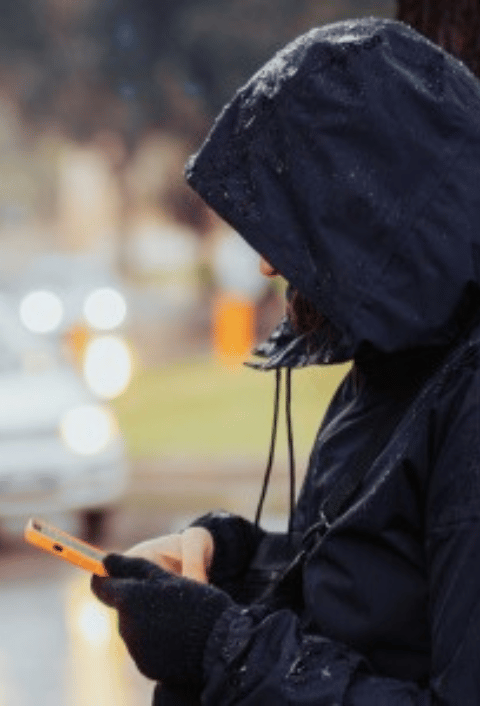 Una persona que lleva una chaqueta impermeable oscura con capucha está de pie al aire libre, mirando hacia abajo a un smartphone. Se ven gotas de lluvia en la chaqueta. El fondo está desenfocado y muestra algo de vegetación, un coche blanco y luces con efecto bokeh, lo que sugiere un día lluvioso en un entorno urbano.